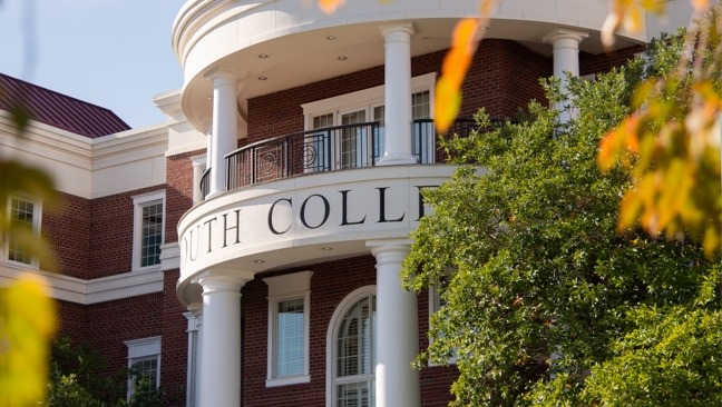 A brick building with white columns and a curved balcony has “SOUTH COLLEGE” partially visible on its facade. The structure is surrounded by trees with leaves showing autumn colors.