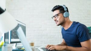 A man wearing glasses and headphones is focused on coding at his desk. He is using a computer with lines of code visible on the screen. His workspace includes a second monitor.