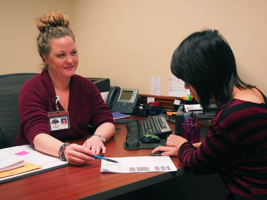 An admissions officer with curly hair, wearing a maroon sweater, sits at a desk discussing admissions paperwork with a student who has dark hair.