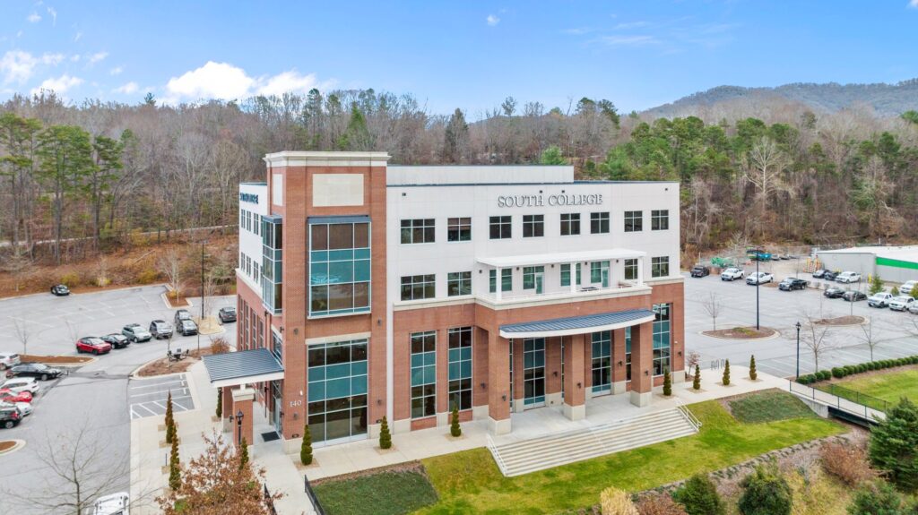 An image showing a large, modern brick building with the words "South College" displayed on the front. The building is surrounded by a parking lot with several cars and is set against a backdrop of trees and hills under a clear blue sky.