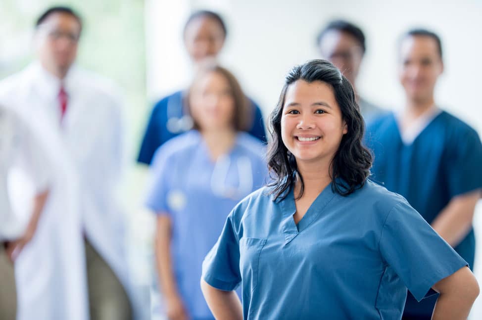 A group of nursing students, both male and female, stand together. The focus is on a woman at the front wearing blue scrubs, smiling confidently.