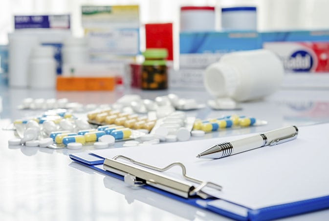 A clipboard with a pen rests on a table surrounded by assorted medication bottles and scattered pills. Blurry boxes of medicine are in the background.