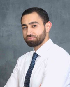 A man with short dark hair, a trimmed beard, and mustache is smiling slightly. He is wearing a white dress shirt and a patterned dark tie. The background is a plain gray textured wall.