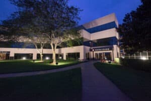 A two-story building of South College, captured during the evening. The building features large windows and is surrounded by a well-maintained lawn and illuminated by pathway lights. The sky is a deep blue, indicating dusk. Trees and bushes are present in the foreground.