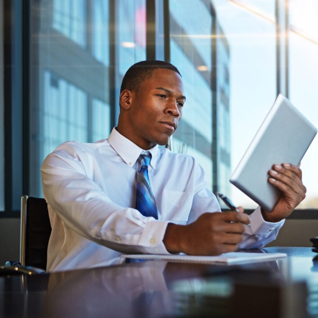 A man in a white dress shirt and blue tie sits at a desk in a modern office, holding a tablet in one hand and using a pen in the other. Large windows in the background reflect the exterior of tall buildings.