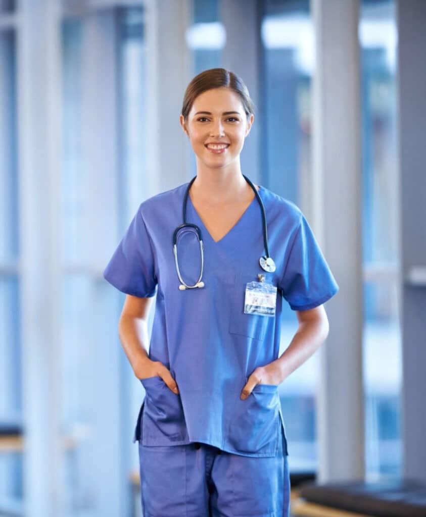 A medical assisting student wearing blue scrubs with a stethoscope around their neck stands in a well-lit medical facility. They have their hands in their pockets and are smiling at the camera. Their ID badge is visible, and large windows are in the background.