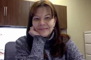 A person with long brown hair, wearing a gray sweater, is seated indoors with their chin resting on their hand. They are smiling gently.