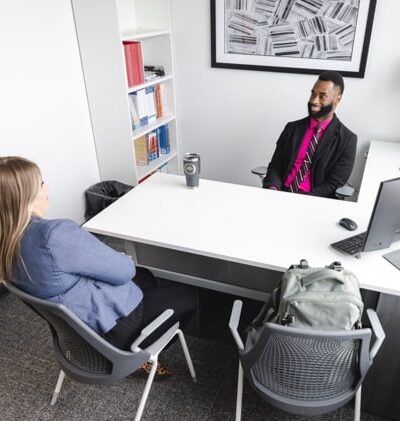 Two people are sitting across from each other in a modern office. One person, wearing a blue jacket, has their back to the camera.