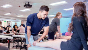 A Physical Therapy student is shown examining a patient's leg in a brightly lit, modern classroom setting. Other students and practitioners are working and observing in the background.