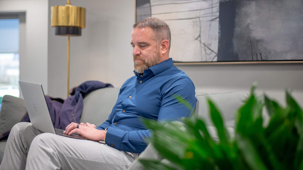 A man with a beard is seated on a gray sofa, working on a laptop. He is wearing a blue shirt and gray pants.