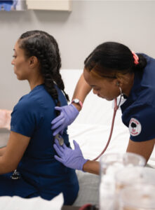 A healthcare student wearing blue scrubs and purple gloves uses a stethoscope to listen to the back of a seated woman in blue scrubs. They are in a clinical setting.