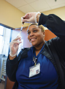 A healthcare student wearing a blue uniform and gloves smiles while preparing a syringe with medication. She stands in a medical facility.