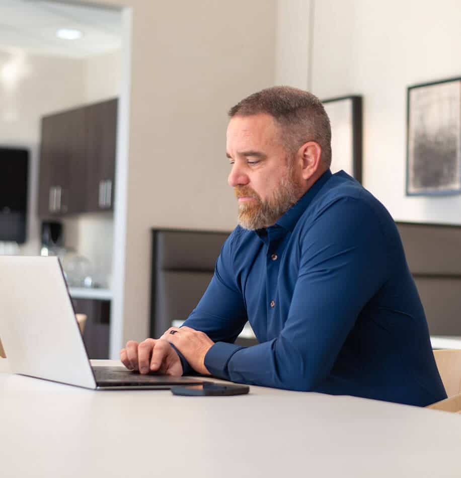 A man with a beard and short hair sits at a table, working on a laptop in a modern office setting. He wears a blue shirt and appears focused on the screen.