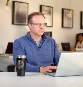 A man with glasses sits at a desk working on a laptop in a modern office. He is wearing a blue sweater and has a coffee tumbler beside him.