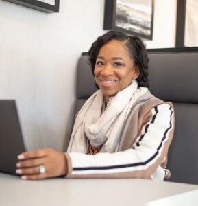 A woman wearing a light-colored scarf and a striped cardigan is sitting at a table with a laptop in front of her. She is smiling.