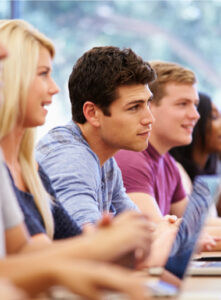 A group of young adults sit at a row of desks in a classroom, attentively facing forward. They are casually dressed, with laptops open in front of them.