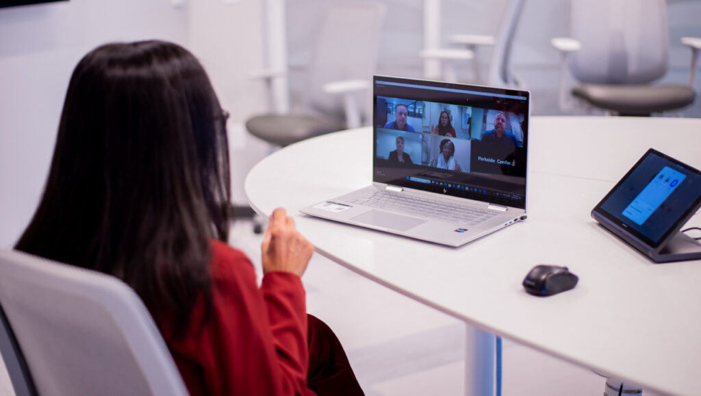 A person with long dark hair in a red shirt sits at a round table participating in a video conference on a laptop.
