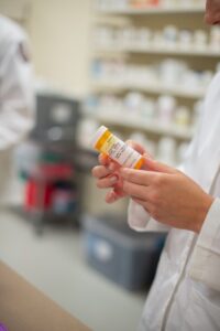 A person in a white lab coat holds a prescription bottle in their hands. Shelves filled with various medications are seen in the blurred background, indicating a pharmacy setting. The person appears to be examining or reading the label on the bottle.