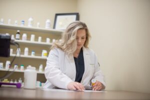 A woman in a white lab coat, with long, wavy blonde hair, is standing and examining a prescription bottle and some paperwork in a pharmacy-like setting. Shelves stacked with various medication bottles are visible in the background.