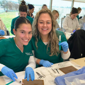 Two smiling medical professionals in green scrubs and blue gloves work together at a table filled with medical tools and syringes in a classroom setting. More individuals in similar attire are focused on their tasks in the background.