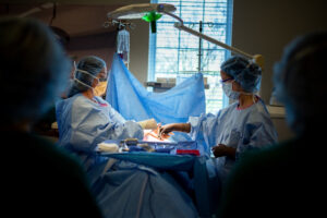 Two surgeons, wearing blue surgical gowns, gloves, masks, and hair covers, operate on a patient in a well-lit operating room. Surgical tools and equipment surround them as light from a window illuminates the scene.