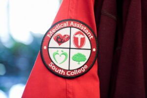 A red medical uniform shows a round patch on the sleeve. The patch reads "Medical Assistant South College" and features four icons: a heart with a stethoscope, a caduceus symbol, heart shape and a tree, representing various health and medical themes.