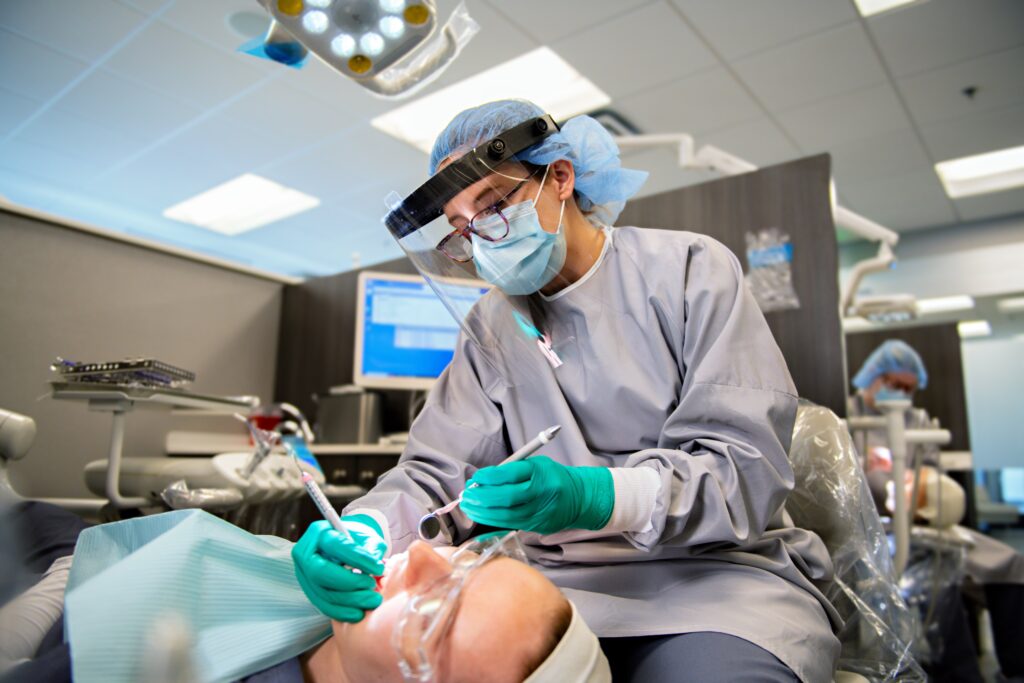 A dental student, wearing a face shield, mask, gloves, and scrubs, performs a dental procedure on a patient. The patient is lying back in the dental chair with mouth open and protective glasses on. The clinic has a modern setup with monitors and dental tools.