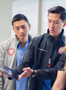 Two healthcare professionals dressed in medical attire, one in a grey jacket and the other in a black one, interact with another person who is partially visible. They appear to be engaged in a discussion, reviewing a tablet.