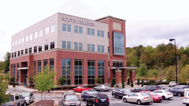 A multi-story building with "South College" signage on the top corner. The structure features large windows and a brick exterior. Several cars are parked in the parking lot in front of the building, and trees are visible in the background.