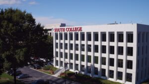 An aerial view of a multi-story white building with the words "South College" prominently displayed in red on the roof. The building has multiple rows of large windows, and a tree and parking lot are visible nearby under a clear blue sky.