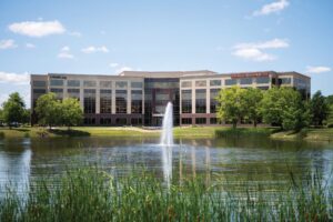 A large campus building with a modern facade sits behind a tranquil pond. A fountain in the middle of the pond sprays water into the air. Trees and greenery surround the water, and the sky above is clear with a few scattered clouds.