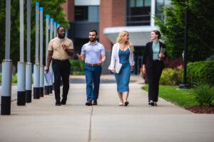 Four people are walking and conversing on a sidewalk. They are in a campus setting, with greenery and a building in the background. Two men and two women are dressed in business casual attire, holding notebooks or folders.