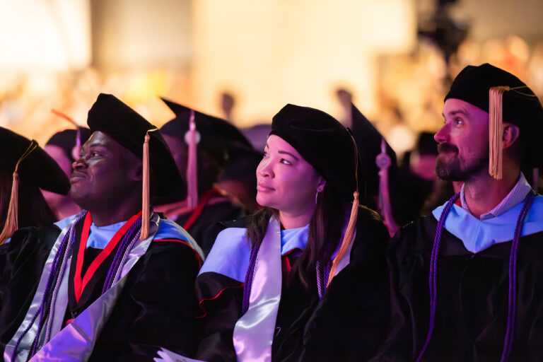 A group of graduates dressed in black academic gowns and caps with tassels are seated and facing forward. They appear to be attending a graduation ceremony, with one man smiling, a woman looking towards the side, and another man looking straight ahead.
