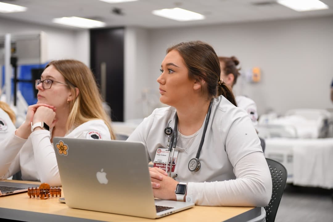 Female Nursing Student sitting in class at desk, with laptop and wearing scrubs and stethoscope