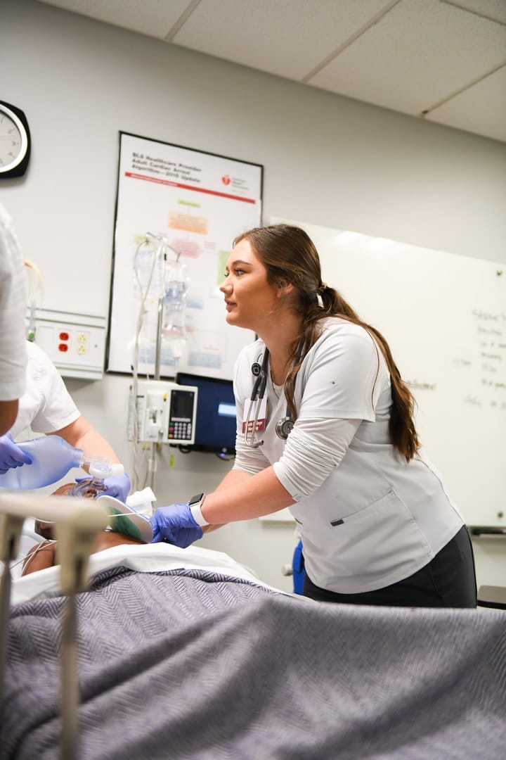 Young female nursing student receiving instruction in mock hospital setting with simulated patient.