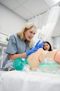 A Respiratory Therapy student practices medical procedures on a training mannequin. She wears blue scrubs and gloves while holding a device over the mannequin. Another student observes in the background. Various medical tools and equipment are visible on the bed.