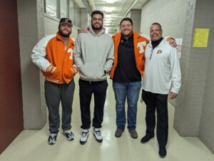 Four men pose together in a hallway. Two are wearing orange varsity jackets, one is in a grey hoodie and dark pants, and the fourth wears a white pullover with a logo on it. They stand close to each other, smiling at the camera. The hallway has grey walls and a white floor.