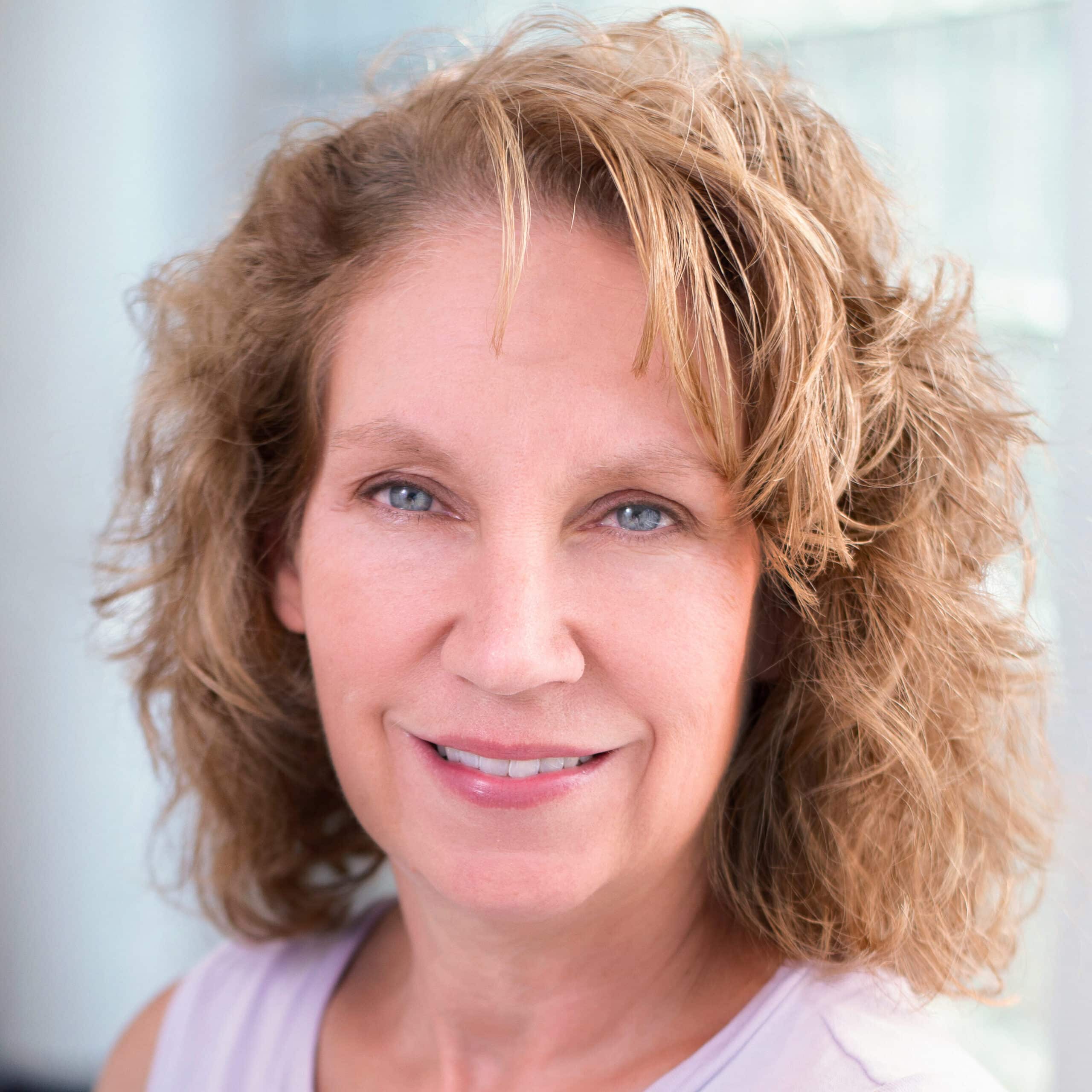 A woman with shoulder-length curly brown hair and blue eyes is smiling at the camera. She is wearing a sleeveless light purple top and has a neutral background with soft lighting.