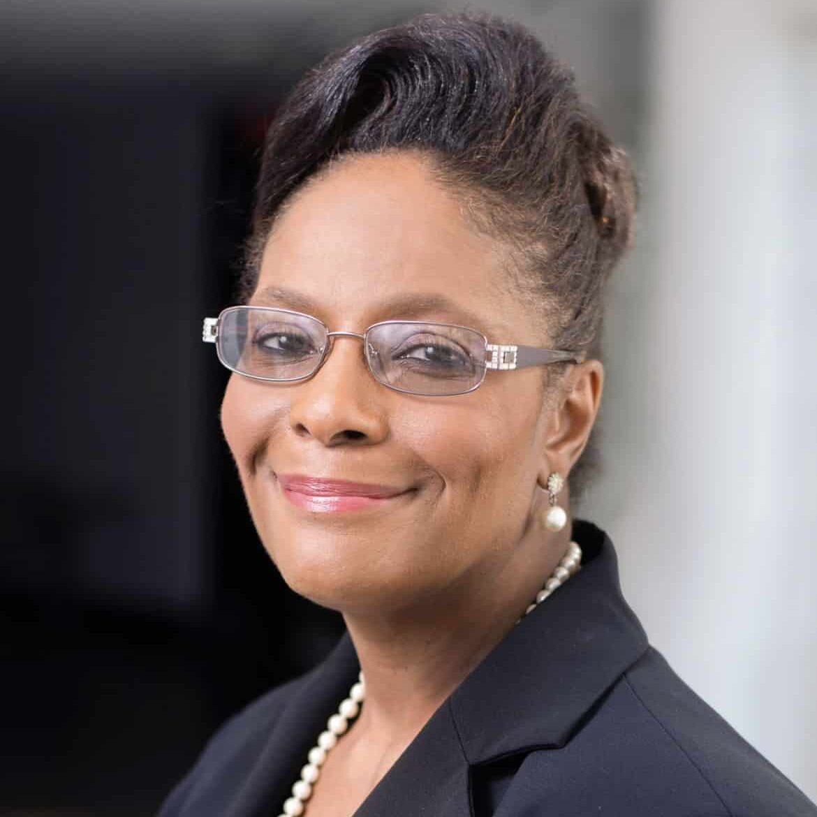 A confident woman with short, styled hair is wearing glasses, pearl earrings, a pearl necklace, and a dark blazer over a patterned top. She smiles warmly at the camera with a professional, blurred background behind her.