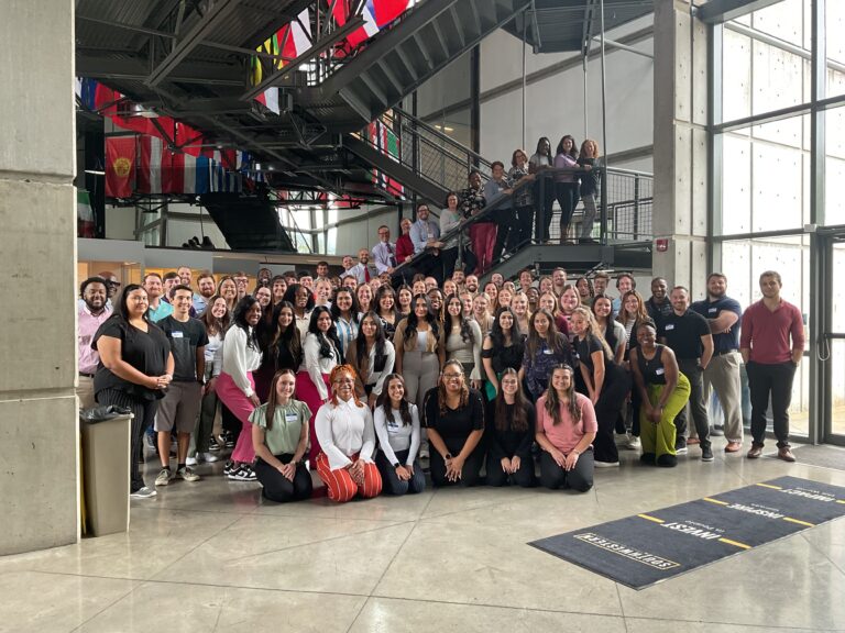 A large group of people poses for a photo inside a modern building with flags hanging from the ceiling. They are standing on multiple levels, including a staircase and the ground floor.