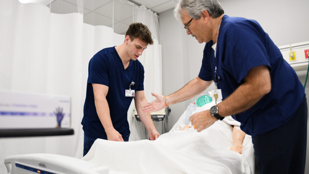 One nursing student and one nursing instructor in navy scrubs assist a patient in a hospital bed.