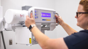 A Radiography student adjusts the controls on an X-ray machine. The technician is wearing glasses and has short hair.
