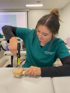 A person wearing a green medical scrub top and black sleeves is practicing inserting a catheter into a simulation model. They are sitting at a table with medical equipment around and a projection screen displaying a presentation in the background.