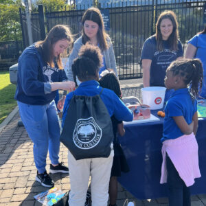 College students in blue shirts and scrubs engage with young children at an outdoor event. They are standing around a table with educational materials and a model of human organs, likely teaching about health or science. One student assists a child closely.