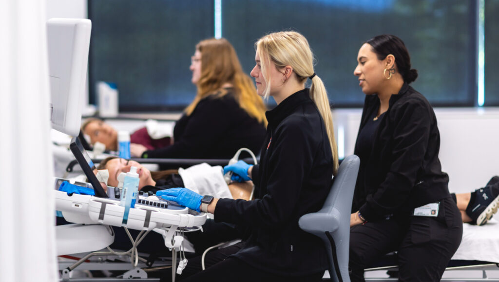 Four people are shown in a medical setting. Two healthcare students are operating ultrasound machines on two patients who are lying on examination beds. The professionals are focused on the monitors, wearing gloves and black clothing.