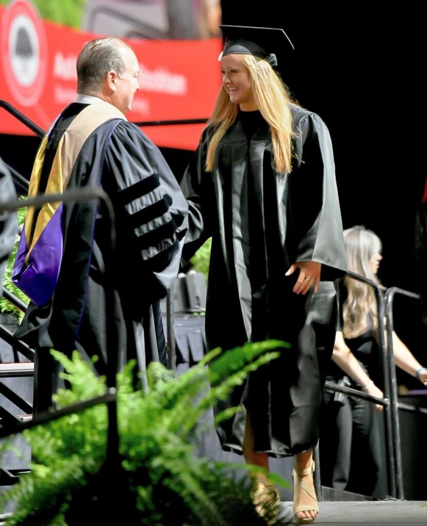 A graduate in a black cap and gown shakes hands with a faculty member on stage during a commencement ceremony. A large screen in the background displays a blurred image. Ferns decorate the front of the stage.