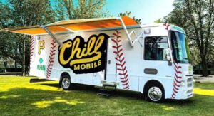 A white food truck labeled "Chill Mobile" with a baseball design and a large yellow awning is parked on grass. It has red stitching patterns resembling a baseball and is surrounded by trees.