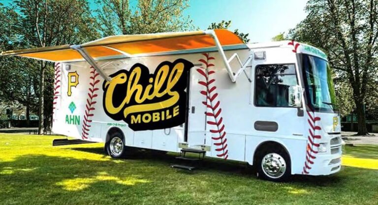 A white food truck labeled "Chill Mobile" with a baseball design and a large yellow awning is parked on grass. It has red stitching patterns resembling a baseball and is surrounded by trees.