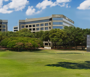 Office buildings with reflective glass windows are surrounded by trees under a blue sky with clouds. Signage for "South College" is displayed on the right side of the building.