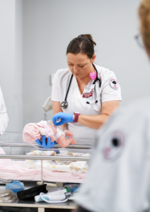A student nurse wearing a white uniform and blue gloves is caring for a newborn in a hospital bassinet. The baby is wrapped in a pink blanket. Medical equipment is visible around them.